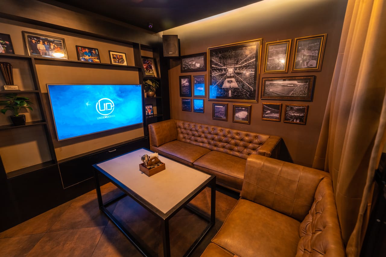A private karaoke room at Underdoggs Aerocity with a leather sofa, a low table, a large screen and framed photographs.