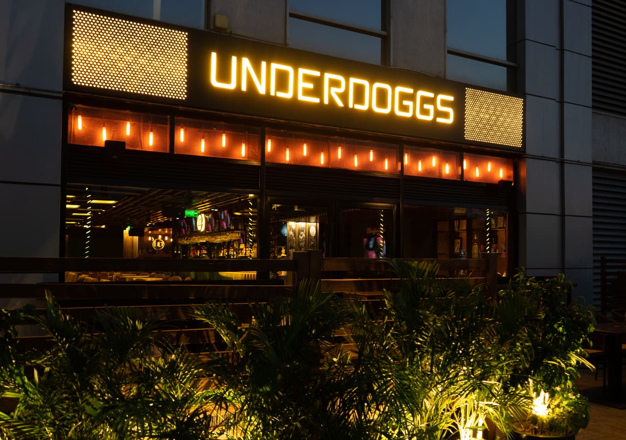 The UNDERDOGGS sign lit above the planted entrance of Underdoggs Cyber Hub at dusk.
