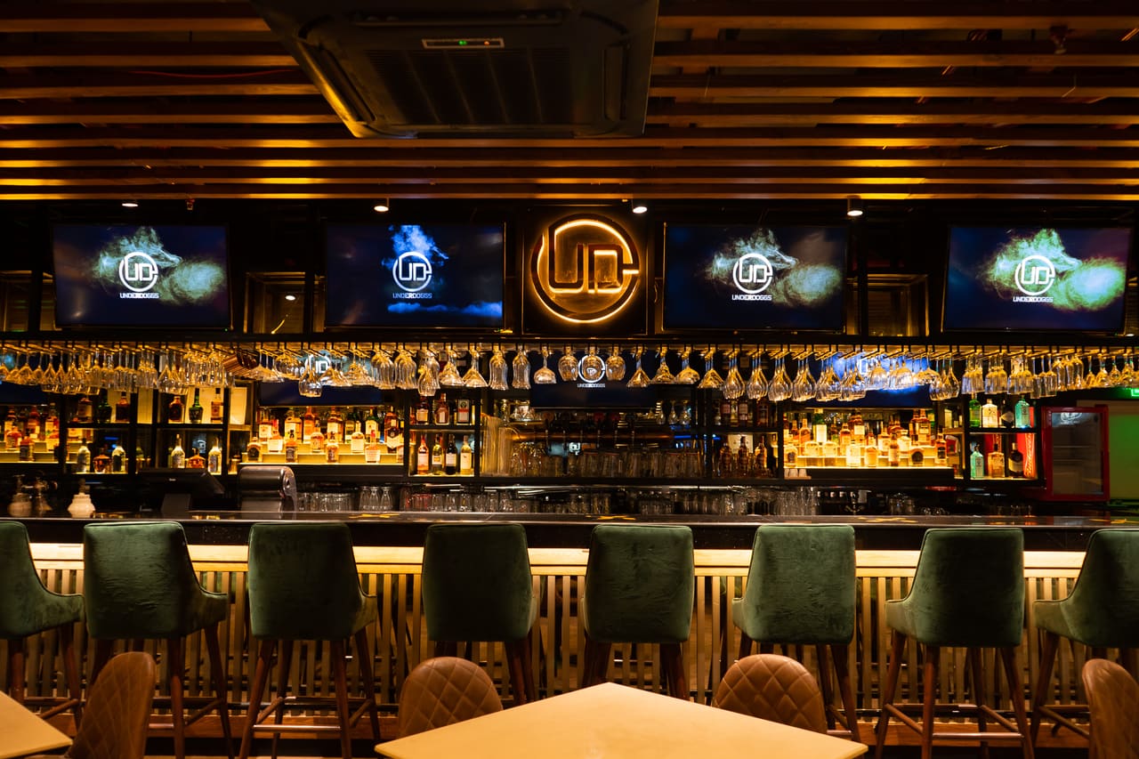 A bar counter with green upholstered stools beneath a backlit UD logo, bottle shelving and screens at Underdoggs Cyber Hub.
